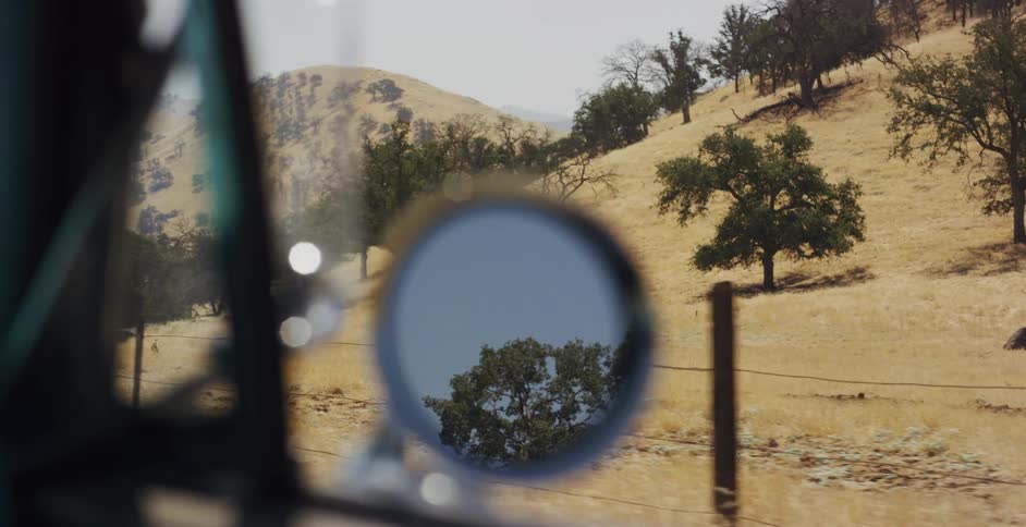 A slow-motion point-of-view shot from a moving car captures the dry, golden hills of the countryside passing by the window