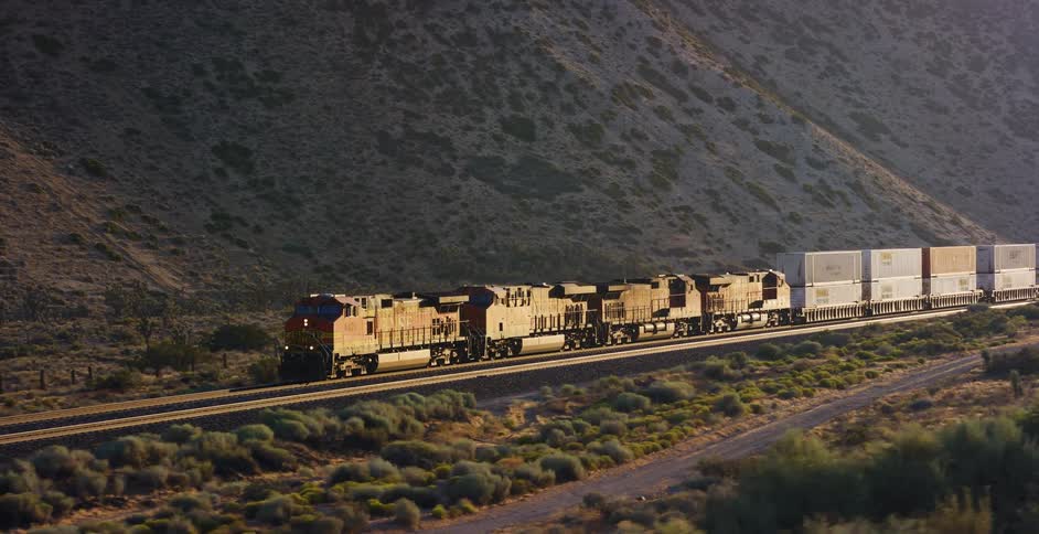 A long freight train hauling shipping containers travels through a vast desert landscape at the base of a mountain during golden hour