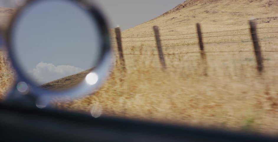 An artistic shot from a moving car shows a fence and dry, rolling hills passing by, with the side mirror reflecting the sky