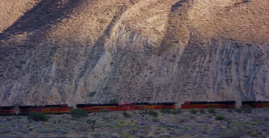 A long freight train travels through a desert landscape at the base of a sunlit mountain during golden hour