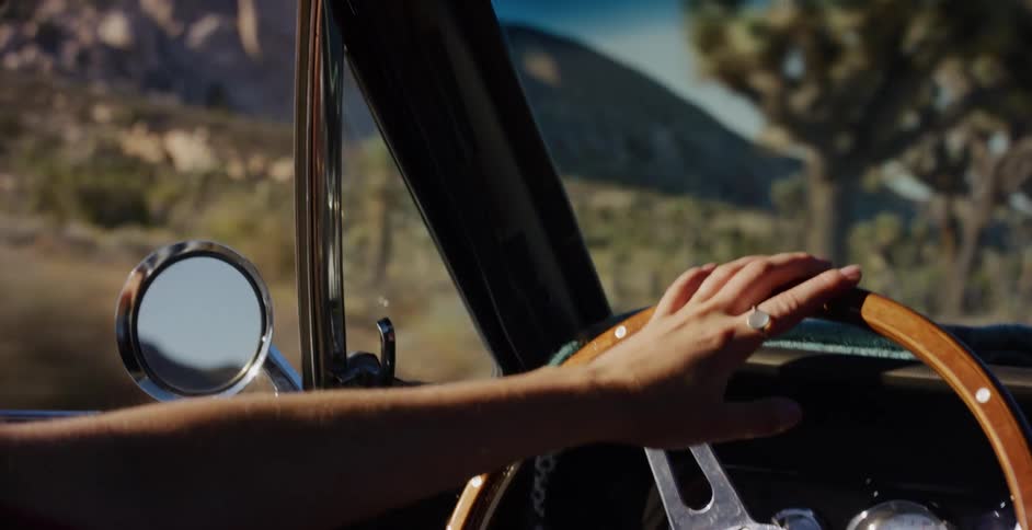 A close-up shot of a person's hand on the steering wheel of a classic car driving through a desert landscape with Joshua trees