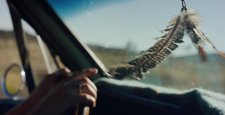 A collection of decorative feathers hangs from a car's rearview mirror, swaying in slow motion during a sunny road trip