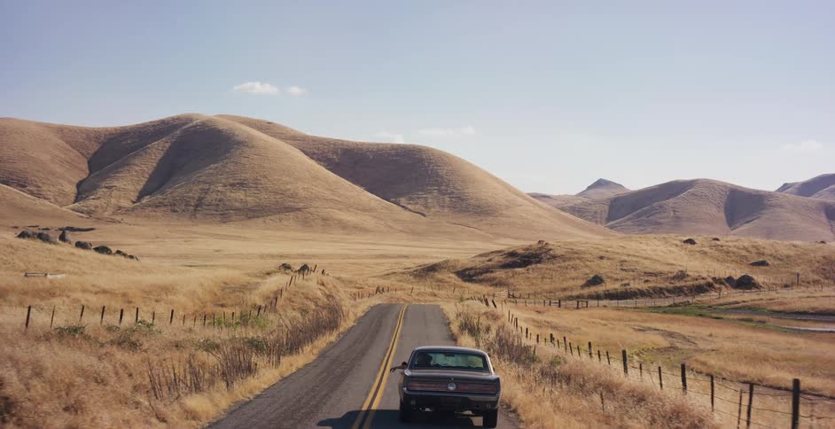Aerial view following a classic muscle car driving down a remote country road through dry, golden rolling hills on a sunny day