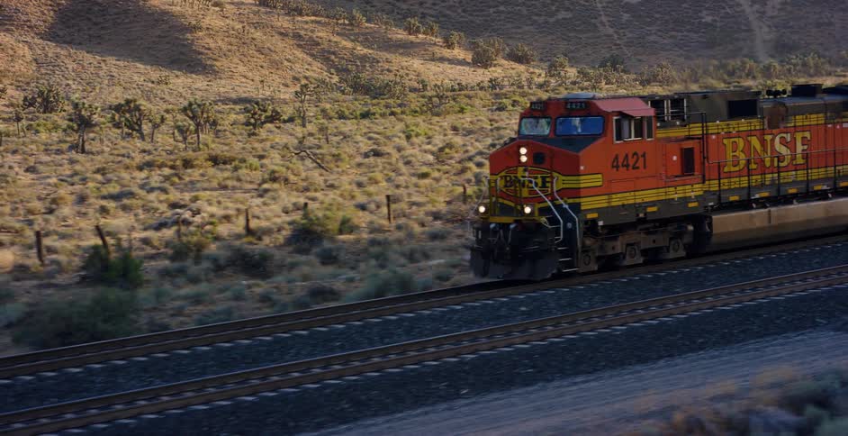 A powerful BNSF freight train travels along railroad tracks through a vast desert landscape dotted with Joshua trees