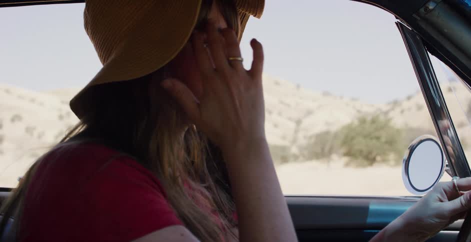A cinematic slow-motion close-up of a thoughtful woman in a yellow hat driving a vintage car through a dry, rural landscape
