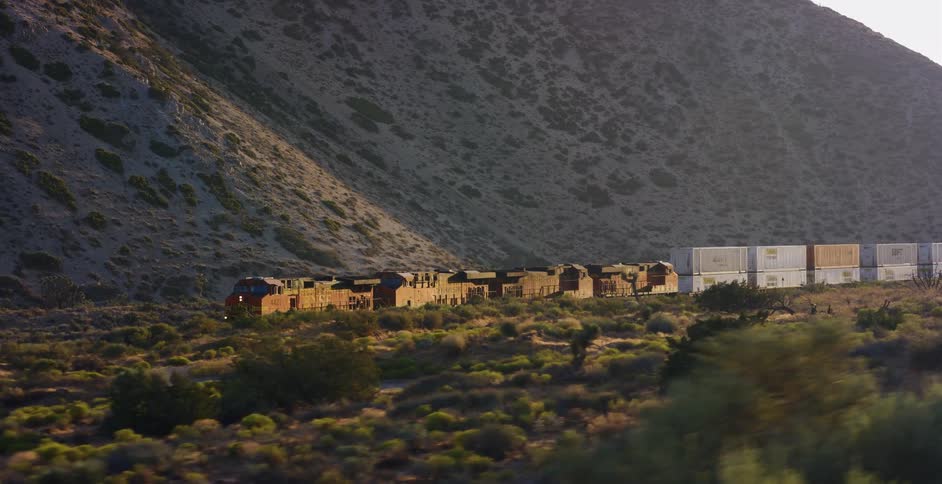 A long freight train travels through a vast desert landscape at the base of a sunlit mountain