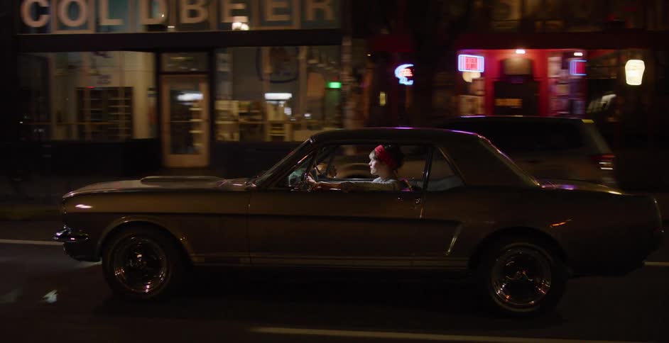 Side profile tracking shot of a stylish woman with a red bandana driving a classic Ford Mustang through the city at night