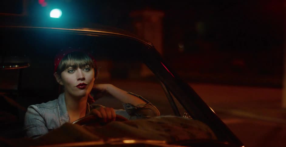 A stylish woman with red lipstick and a bandana sits in her vintage car at a traffic light during a night drive through the city