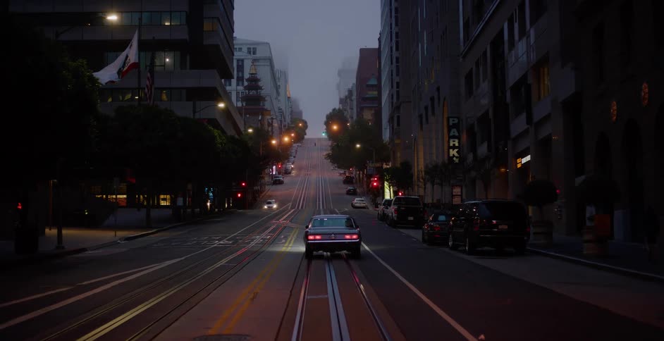 A classic American car drives away up a steep, foggy San Francisco street with cable car tracks at twilight