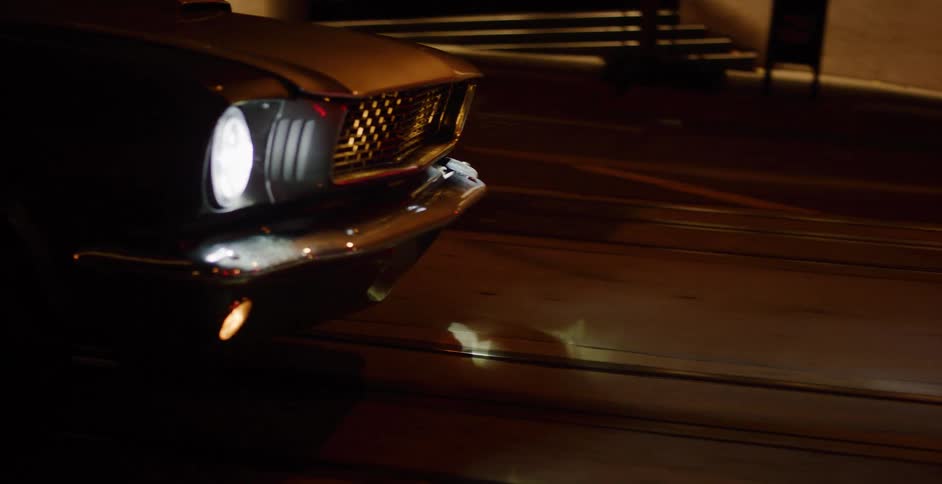 A classic black muscle car cruises down a city street at night, its headlight cutting through the darkness
