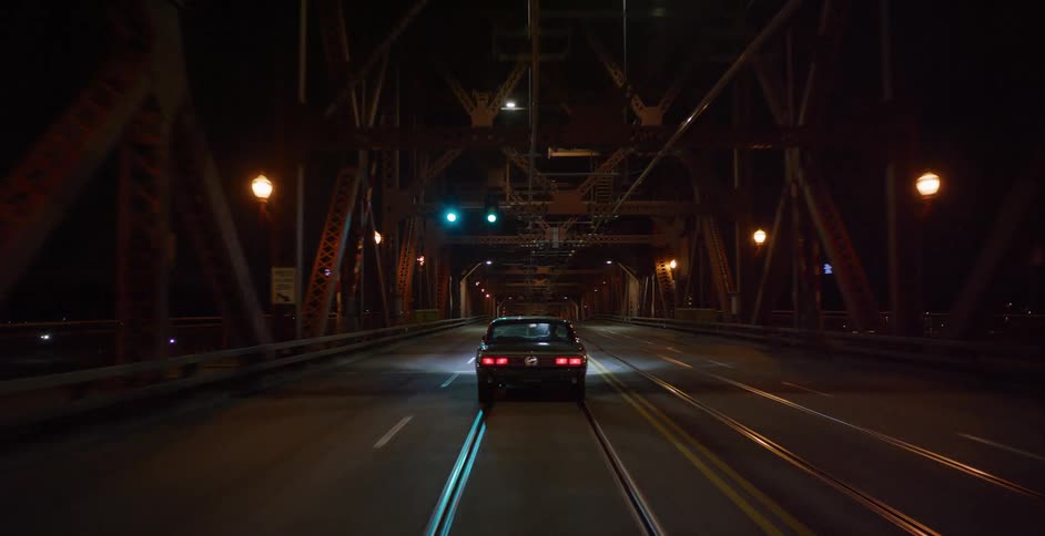 Following a classic dark sedan as it drives over an illuminated steel truss bridge at night in the city