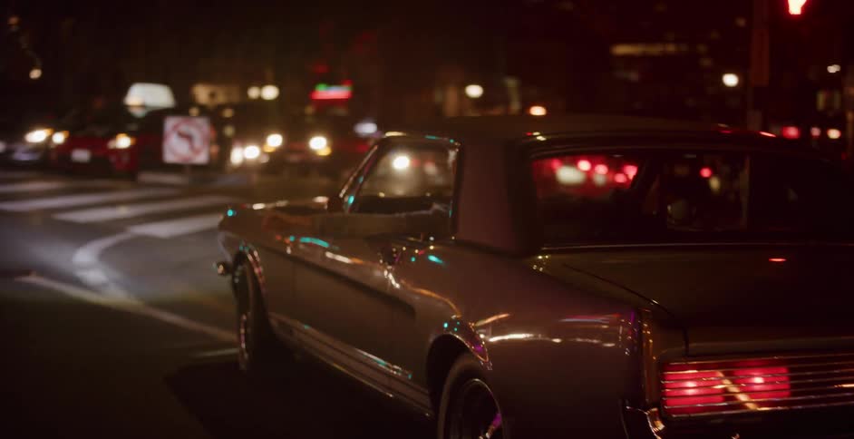 A person with their arm out the window drives a classic muscle car down a neon-lit city street at night