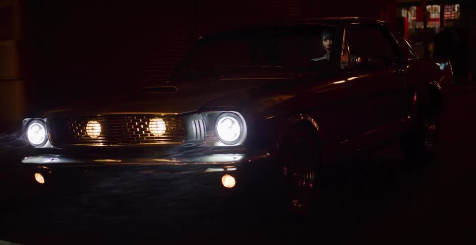 A woman in a vintage muscle car drives through misty city streets with headlights on at night