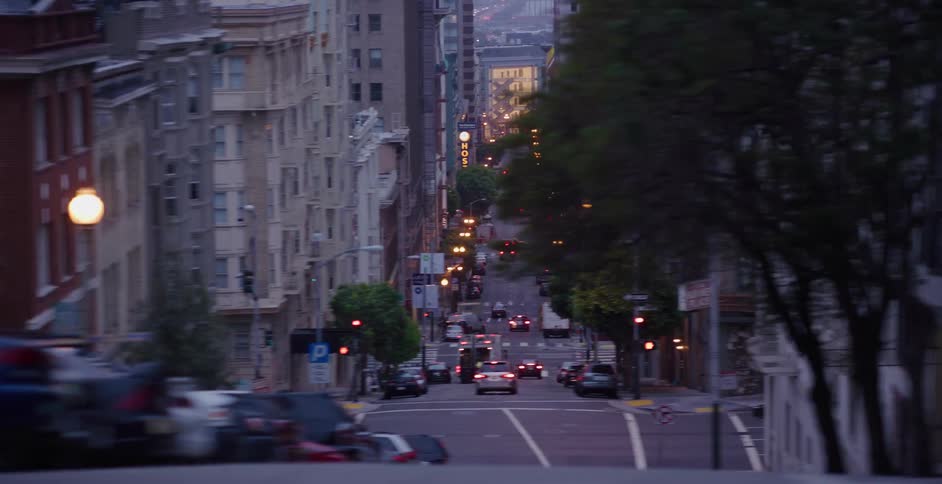 Driving past a steep downtown street filled with cars and illuminated by city lights at twilight