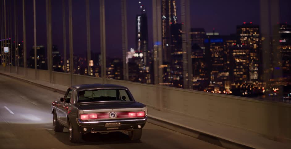 Rear view of a classic muscle car driving on a bridge at night with an illuminated city skyline in the distance
