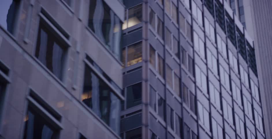 A soft-focus shot pans up a modern office building with glowing windows, revealing a larger skyscraper behind it at twilight