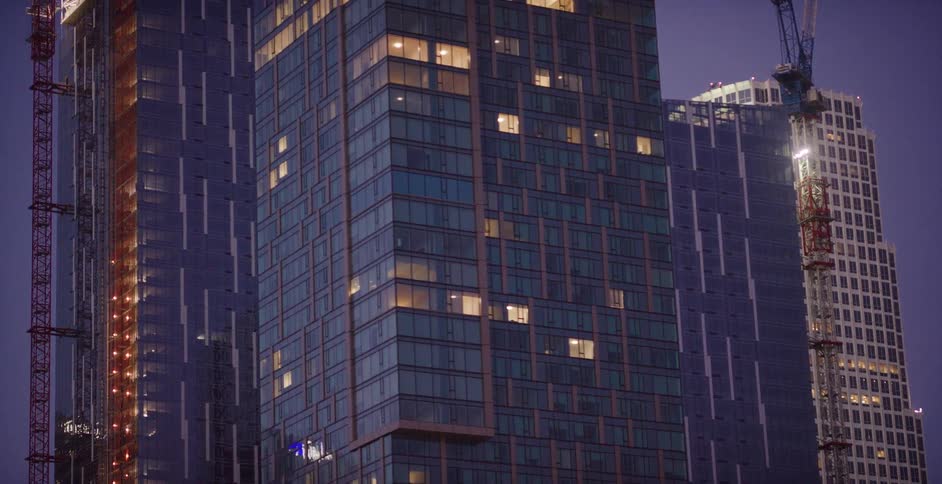 A street lamp flare moves across the glass facade of a modern skyscraper in a developing urban city at dusk