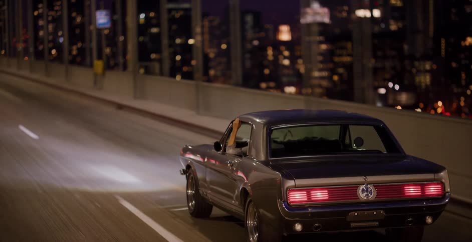 A classic muscle car cruises along a city bridge at night with the beautiful bokeh skyline in the background