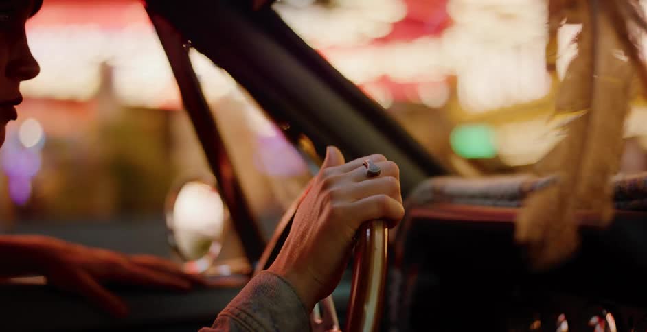 A cinematic close-up shows a woman's hands gripping the wooden steering wheel of a vintage car while driving through a vibrant city at night