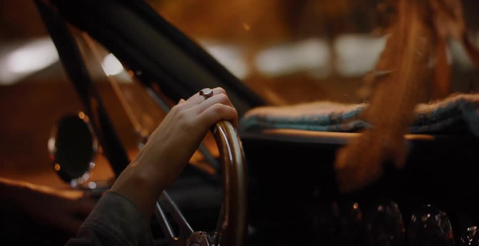 A person's hands grip the wooden steering wheel of a classic car while driving through a vibrant city at night