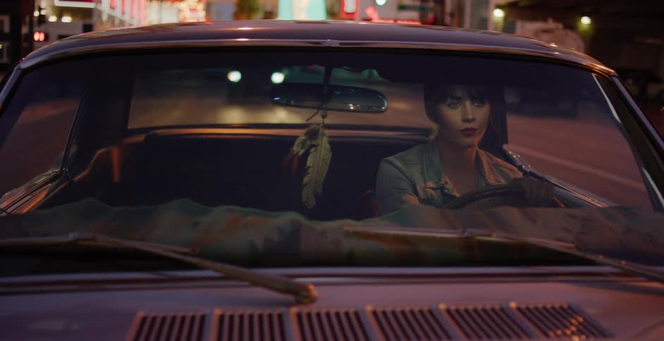 Front view of a young woman driving a vintage car through a neon-lit city street at night