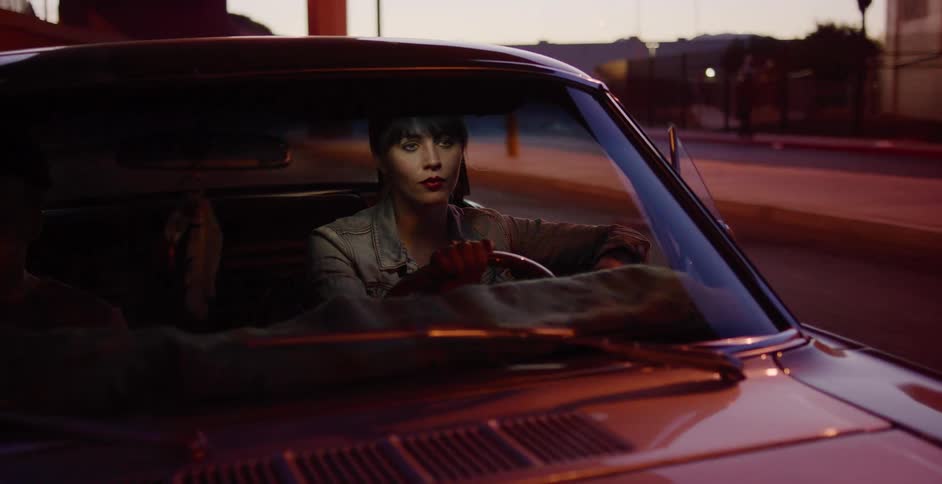 A serious young woman waits behind the wheel of her classic car on a dimly lit city street at dusk