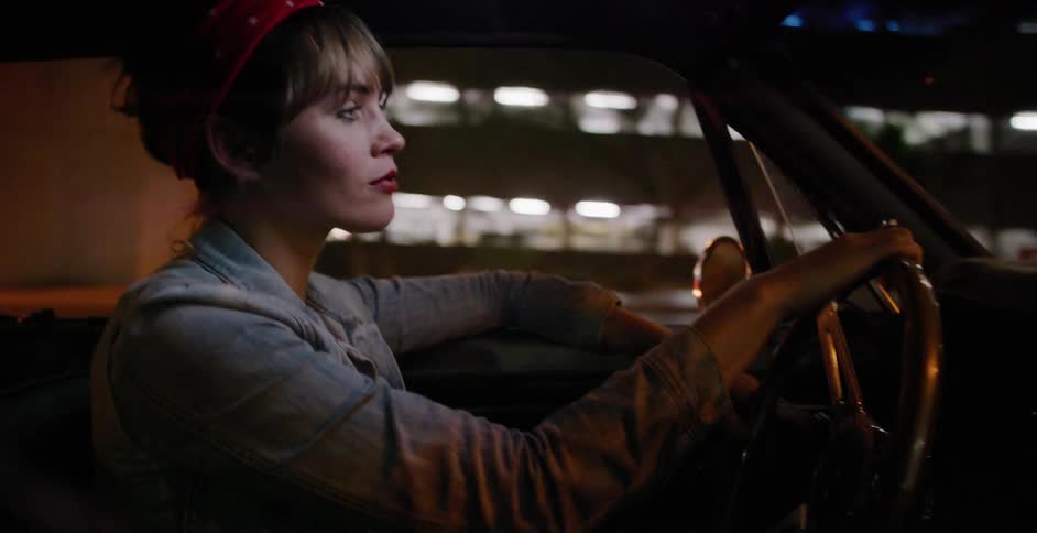 Side profile of a young woman in a denim jacket driving a classic convertible through the city at night
