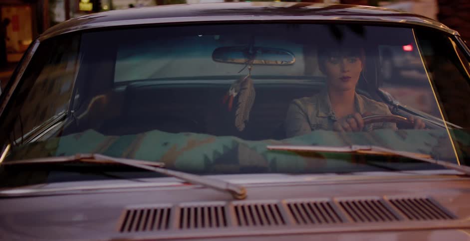 A young woman with short hair waits in her vintage muscle car at dusk as city lights reflect on the windshield