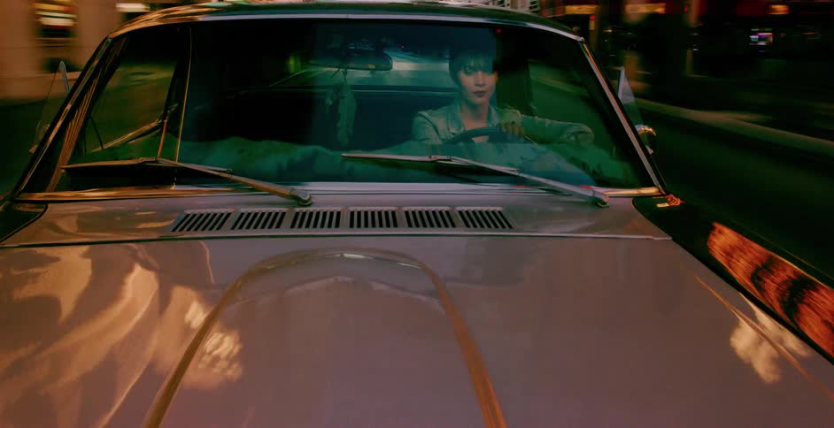 Hood-mounted camera view of a woman driving a vintage car through a neon-lit city at night