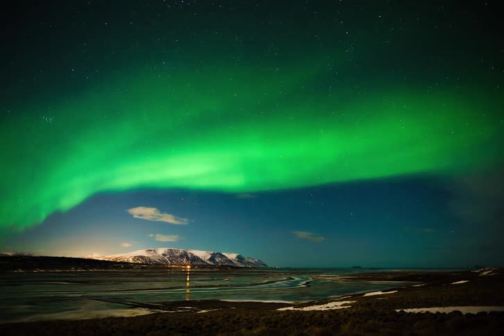 A stunning time-lapse of the vibrant green Aurora Borealis dancing across the starry night sky over a snowy mountain landscape and a frozen lake