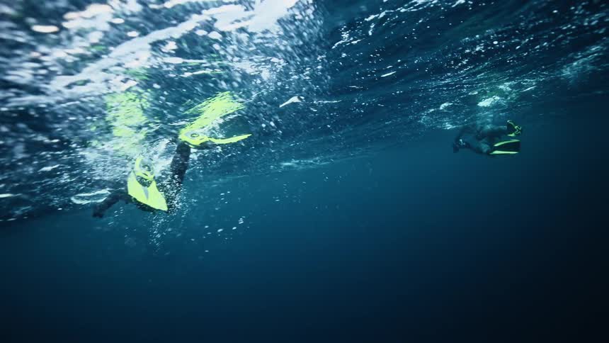 From beneath the waves, two snorkelers with bright yellow fins glide across the surface of the deep blue ocean