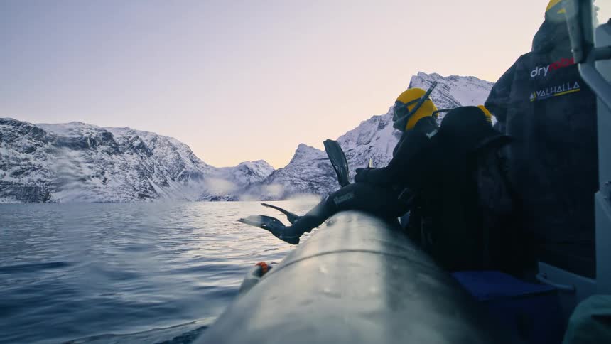 A diver in a full wetsuit sits on the side of a moving inflatable boat, preparing for a swim in a cold fjord surrounded by snowy mountains