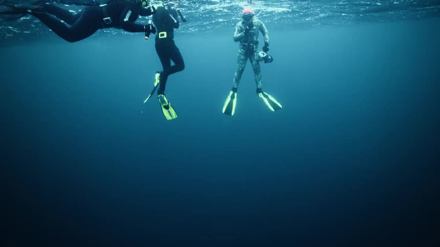 Three divers in full gear float at the surface of the deep blue ocean in slow motion, as seen from below