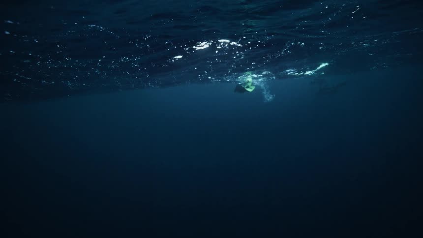 An underwater view captures two scuba divers floating on the dark, rippling surface of the deep ocean