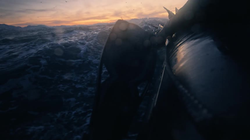 A slow-motion shot from an inflatable boat shows divers in wetsuits navigating choppy ocean waters during a dramatic sunset