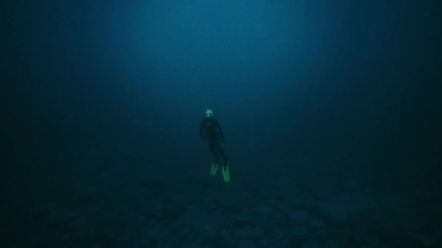 A solitary scuba diver explores the deep, dark ocean floor, turning to face and wave at the camera