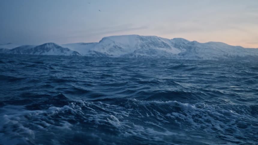 Dramatic slow-motion view of the powerful, dark blue waves of a frigid sea, with snow-capped mountains visible on the horizon