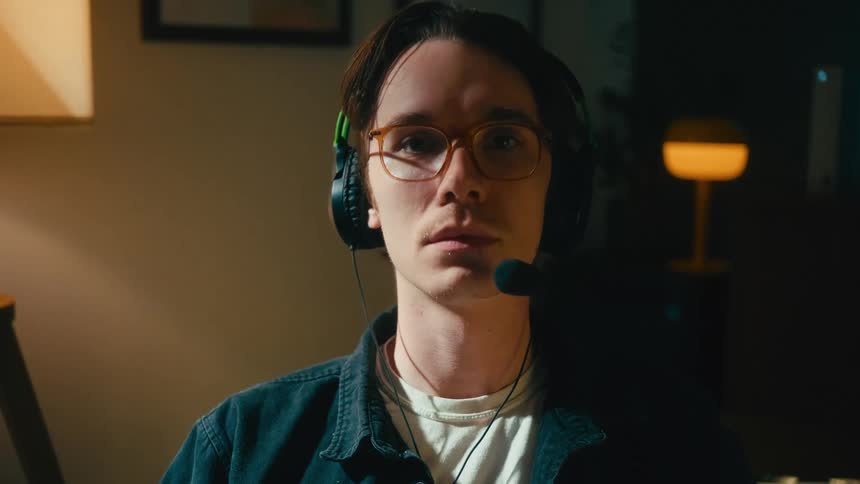 Close-up portrait of a serious young man wearing a gaming headset and glasses, looking intently into the camera in a dimly lit room