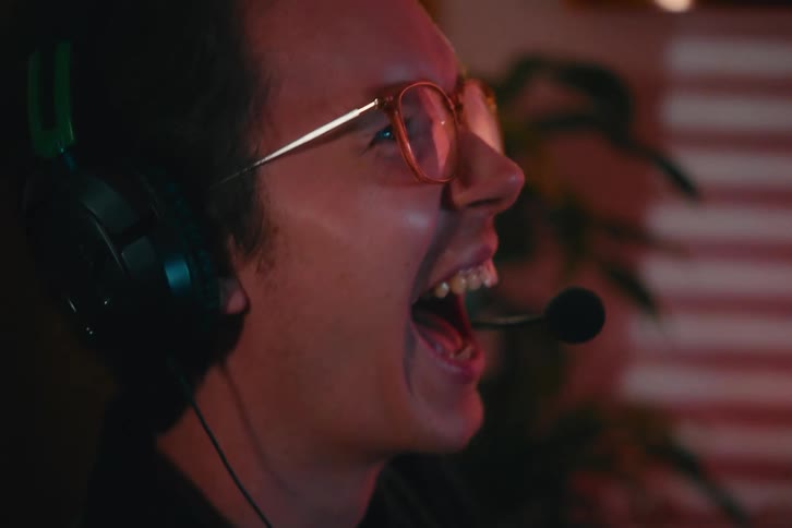 An overjoyed gamer wearing glasses and a headset screams with excitement and joy while playing a video game at night