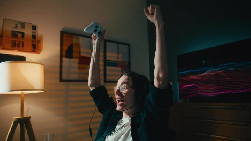 Triumphant young gamer raises his controller in the air and celebrates winning a video game in his cozy room at night