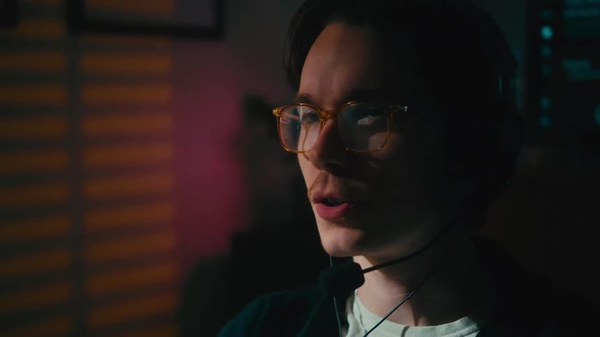 A young man with glasses and a headset talks into a microphone while gaming online at night