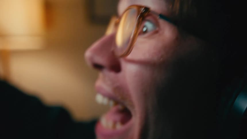 A shaky, extreme close-up of a man with glasses yelling with intense excitement in a dimly lit room at night