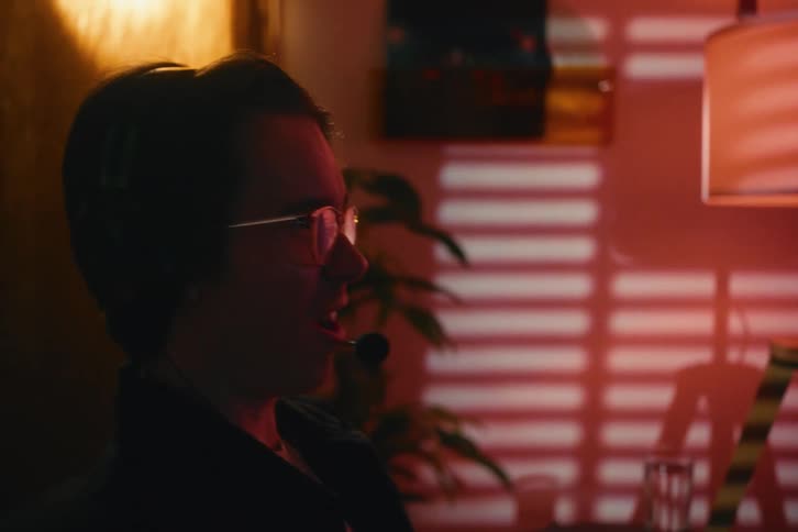 A focused young man with a prosthetic arm wears a headset and talks animatedly while playing a video game in a moodily lit room