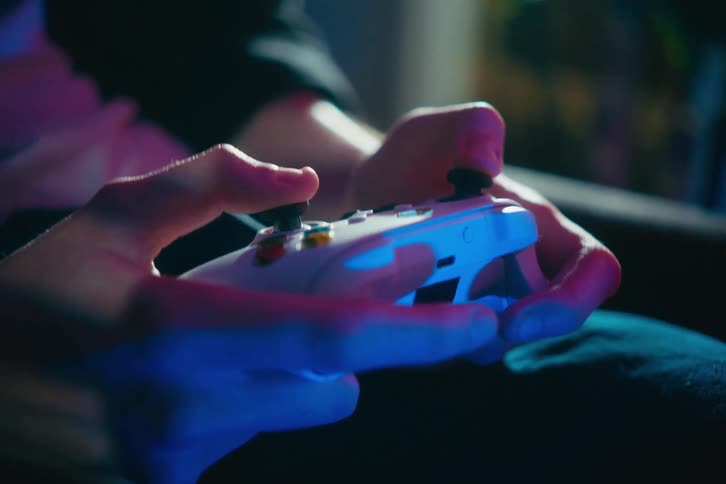 Close-up on a person's hands holding a white gaming controller and playing a video game in a dark room with blue and pink mood lighting