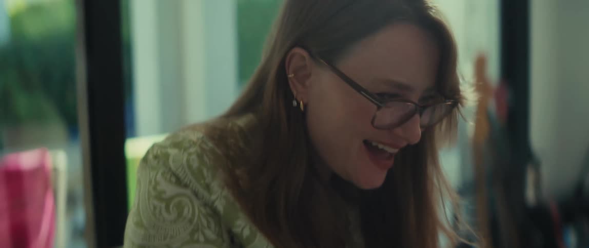 A close-up shot of a smiling woman in glasses happily working on her laptop and taking notes in her journal at a glass desk