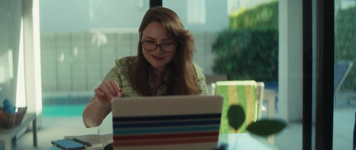 A smiling woman puts on her glasses and works on her laptop at a desk with a view of a sunny backyard