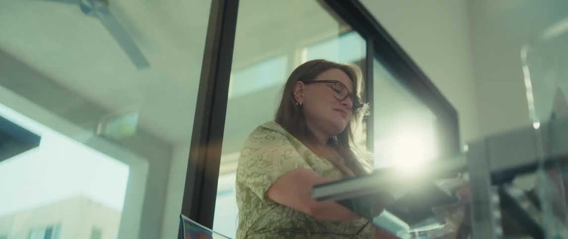 A low-angle view of a woman in glasses meticulously scanning a book in her sun-drenched office