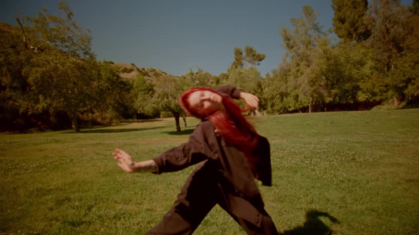 A powerful woman with long, bright red hair practices a slow-motion, expressive martial arts routine in a sunlit park