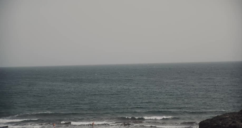 Distant swimmers brave the choppy grey ocean on an overcast day at a rocky beach