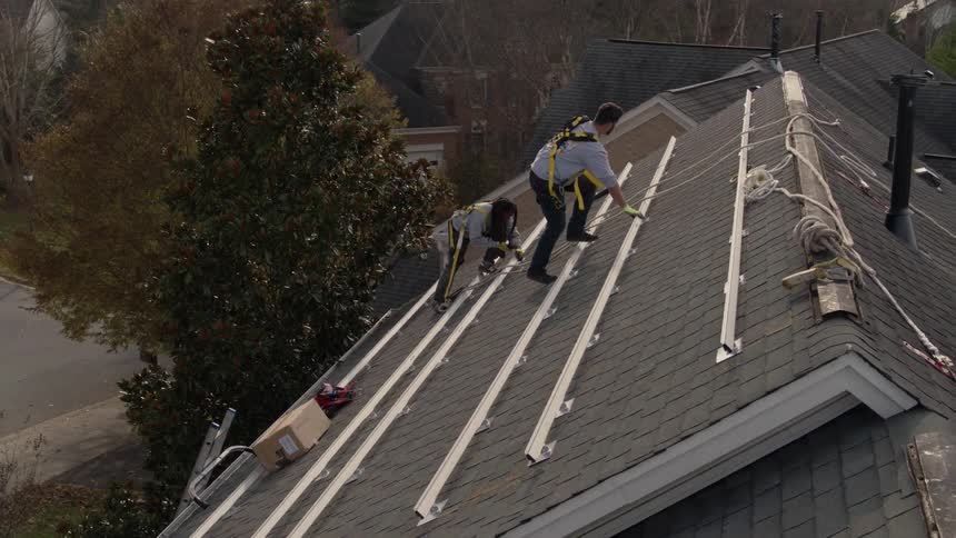 Two men wearing harnesses are installing solar panels on a house